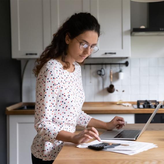 Female standing in home kitchen doing accounting work from home, use calculator fill data results on laptop application