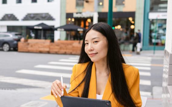 Young Chinese female with credit card and tablet looking away and thinking about online purchases while sitting at table in street cafeteria in Bay Area