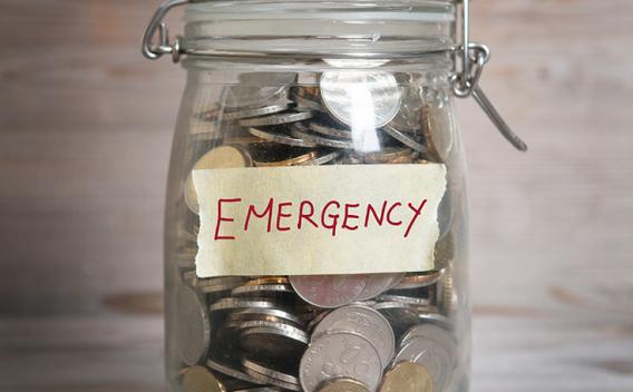 Coins in glass money jar with emergency label, financial concept. Vintage wooden background with dramatic light.