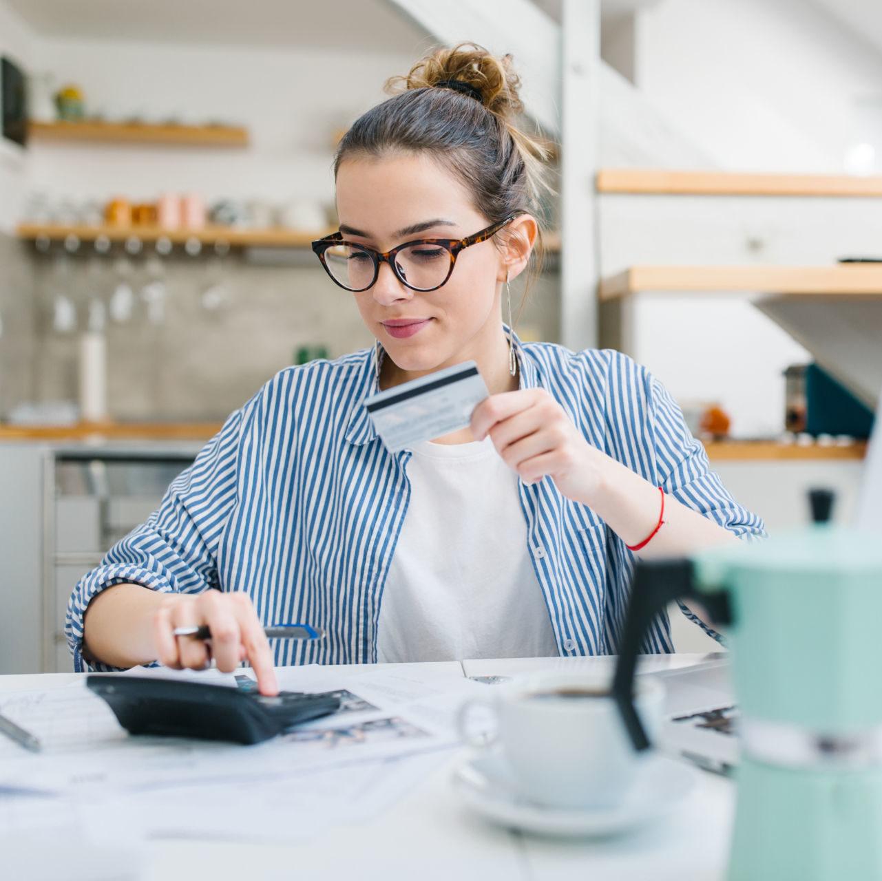 A young woman holding a credit card and using a calculator while sitting at a kitchen table