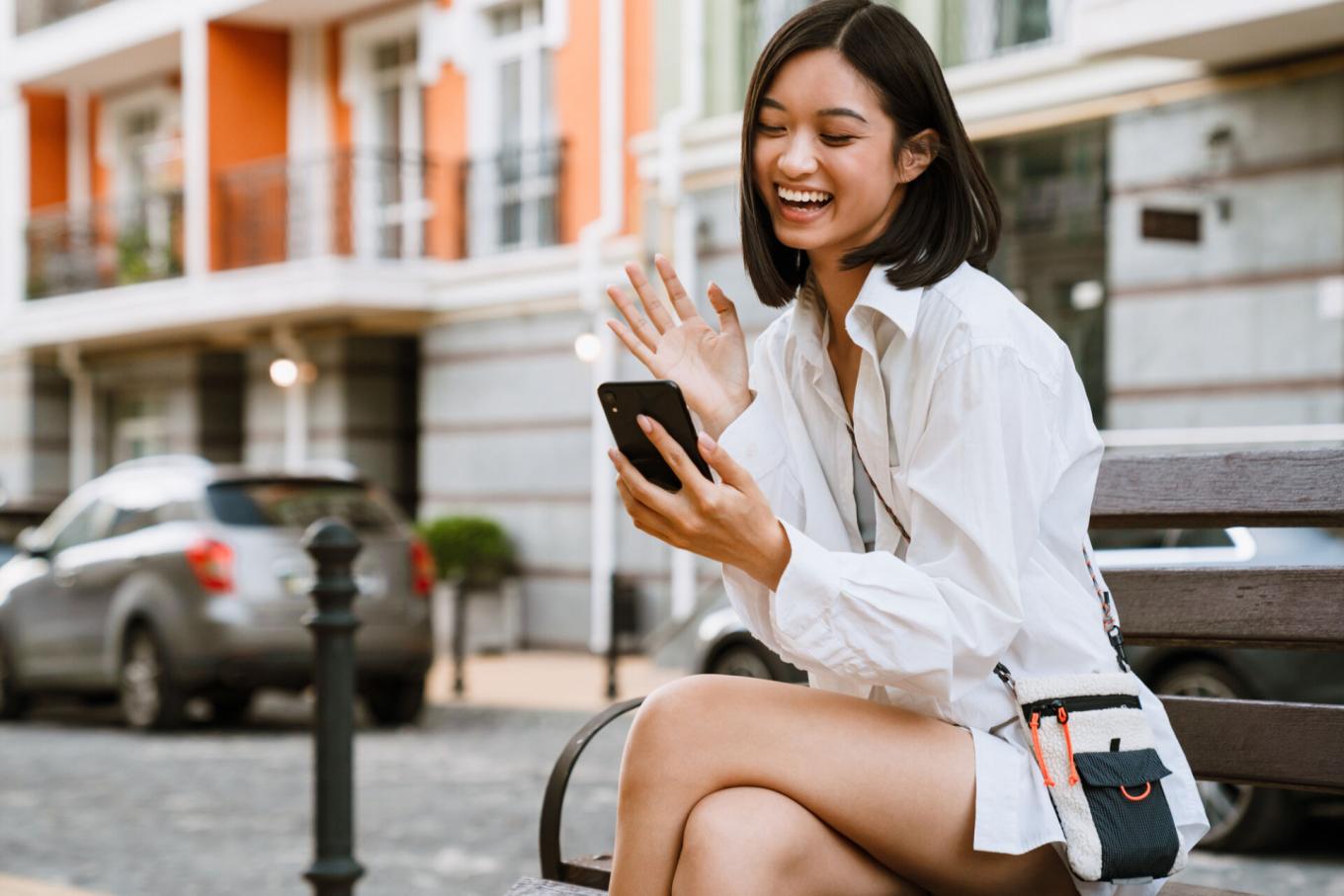 Woman sitting on a bench outside, laughing and waving while looking at her phone