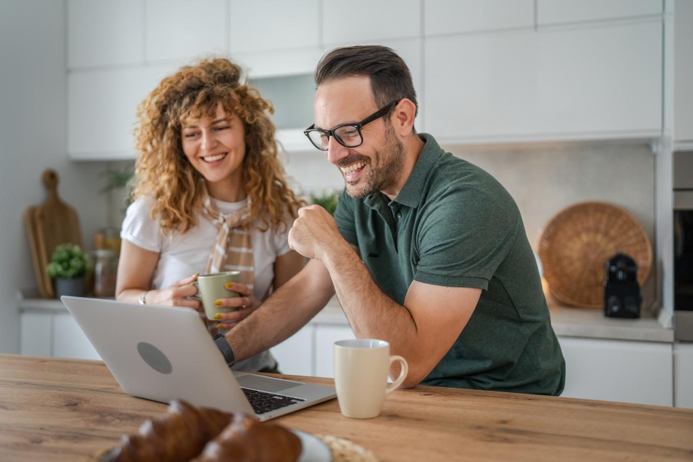 A happy couple drinking coffee in a kitchen while looking at a laptop